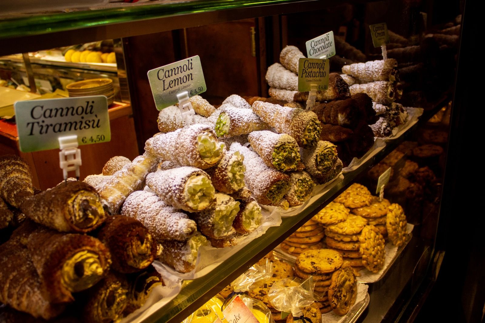 Vitrine de cannoli italiens étiquetés (tiramisù, citron, chocolat, pistache) et de biscuits dans un café.